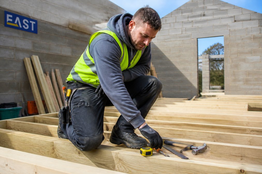 A builder on a construction site wearing a hi-vis vest, reinforced knee work trousers with holster pockets, and steel toe cap boots, laying out tools on a timber frame in daylight