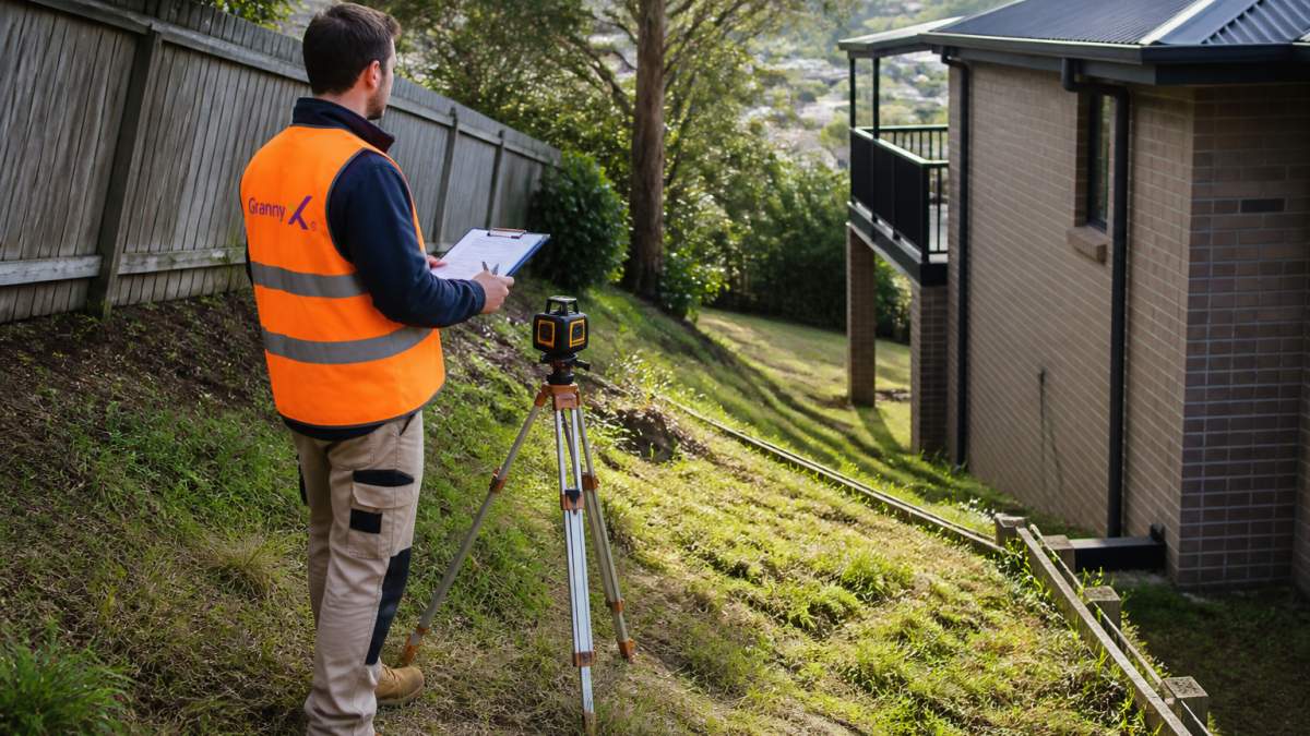 Site assessor evaluating a granny flat sloping block with a laser level and clipboard in a backyard