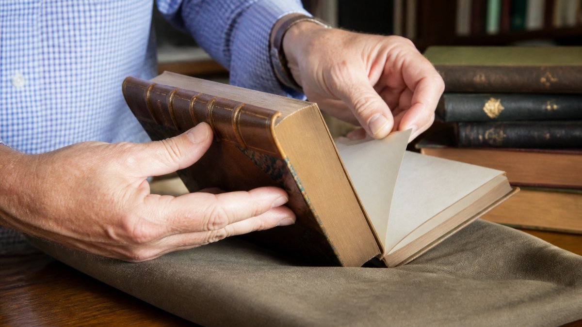 Close-up of hands carefully handling a leather-bound classic novel with a dust jacket, showing proper support of the spine and gentle page turning