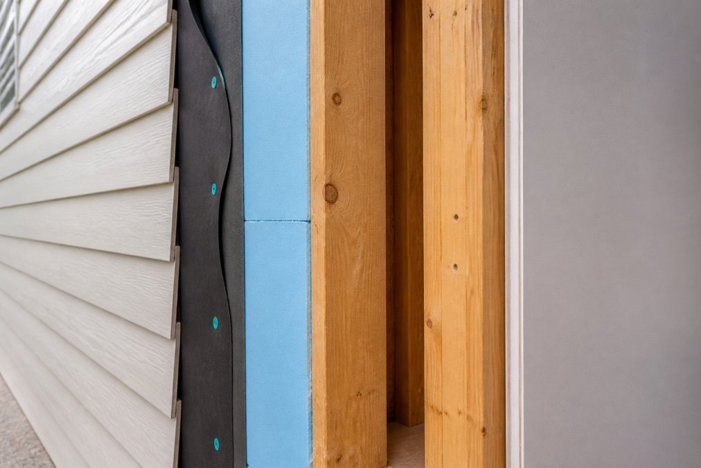 Cross-section view of a home's exterior wall assembly showing visible layers including exterior cladding, weather barrier membrane, rigid foam insulation, wooden framing studs, and interior drywall with natural light highlighting each distinct layer
