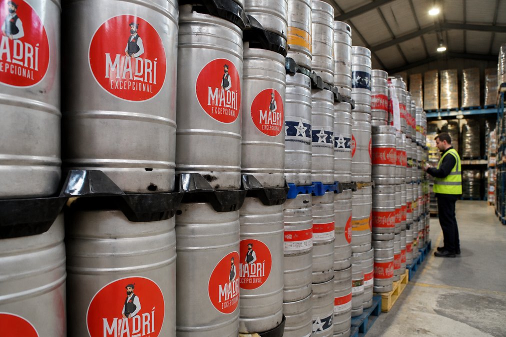 Rows of premium kegs stacked in a warehouse setting, showing different branded keg varieties with distinctive labels under bright warehouse lighting, staff member checking stock in the background