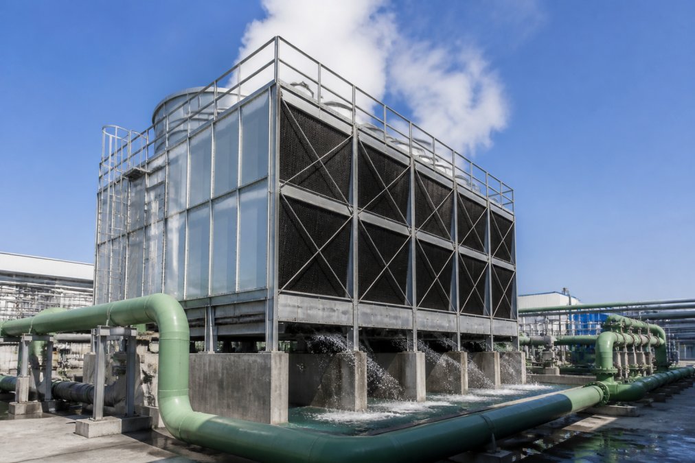 Large industrial cooling tower at an operational facility with pipes and water flow visible, steam rising from the top, shot during daytime with a clear blue sky
