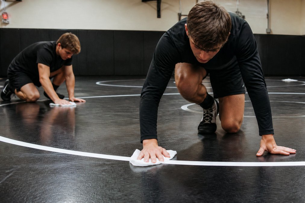 Athletes wiping down wrestling mat with antibacterial wipes during practice, showing the cleaning action and mat surface with natural gym lighting