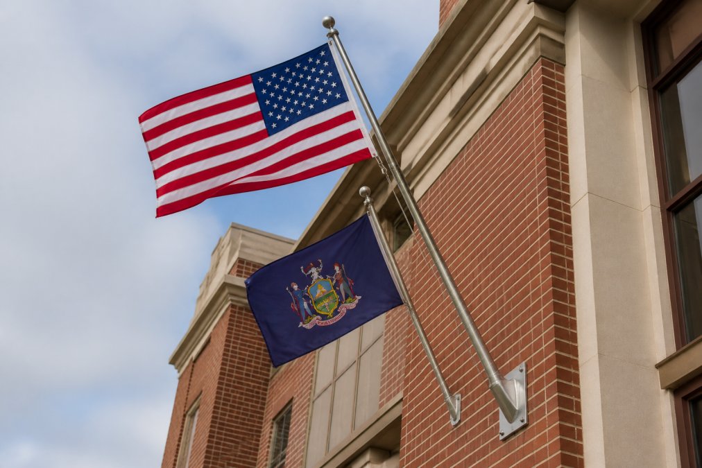 Wall-mounted flagpole system displaying the American flag above a slightly smaller state flag on a brick government building exterior, showing proper hardware installation and positioning