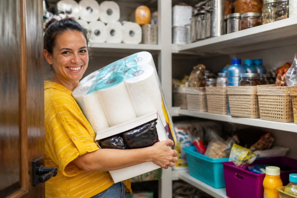 A parent in a bright, organized pantry holding a large pack of paper towels and a box of trash bags, smiling at the camera