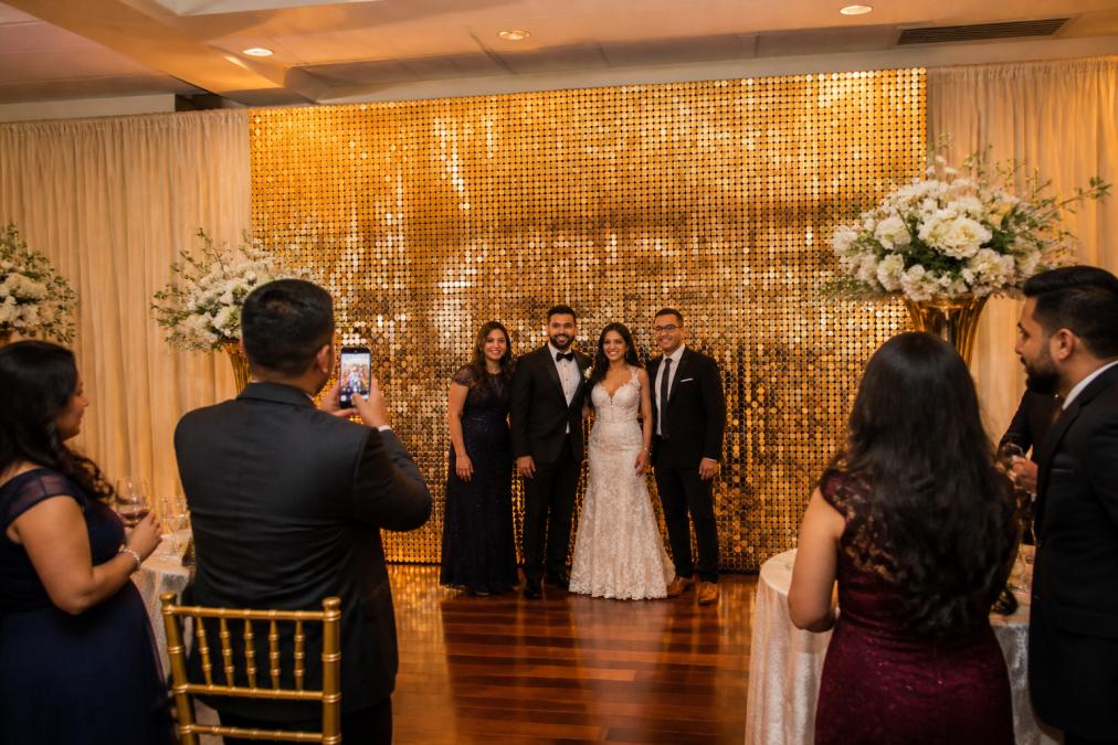 Finished shimmer wall backdrop at an elegant wedding reception with warm professional lighting highlighting the sequin movement and glitter finish, guests in formal attire posing and photographing in front of the backdrop