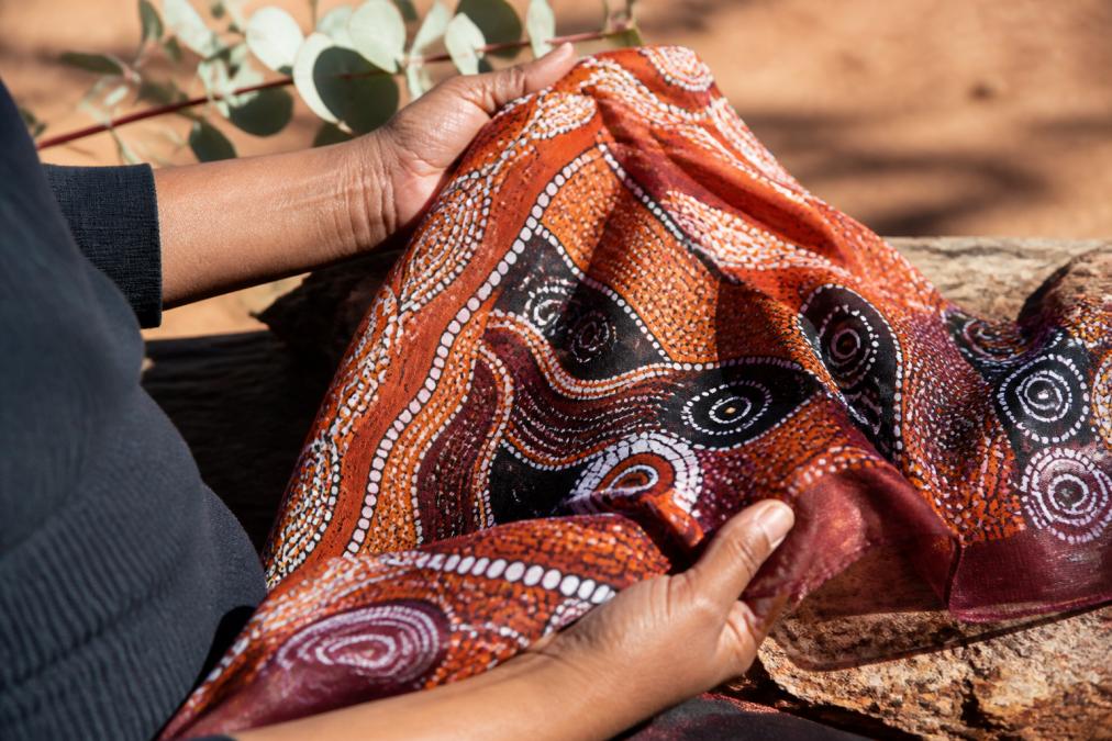 Hands holding a beautifully crafted Indigenous art scarf with intricate traditional patterns, natural sunlight highlighting the vibrant colours and fine textile details