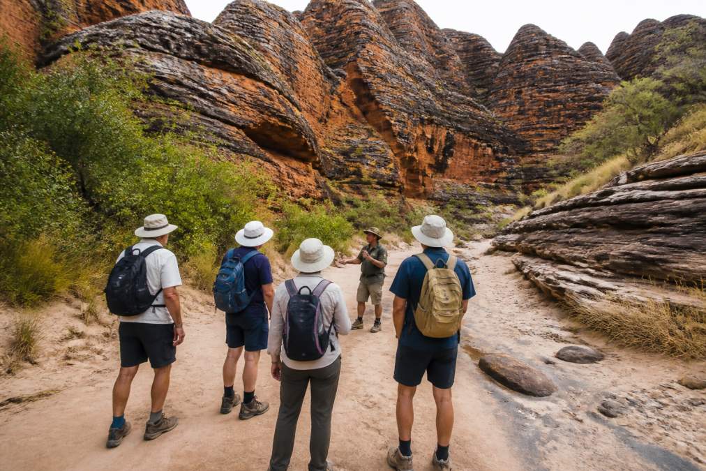 A small group of mature walkers in hats and daypacks moving slowly along a sandy creek bed between towering orange and black striped sandstone domes, with a guide gesturing toward the rock formations