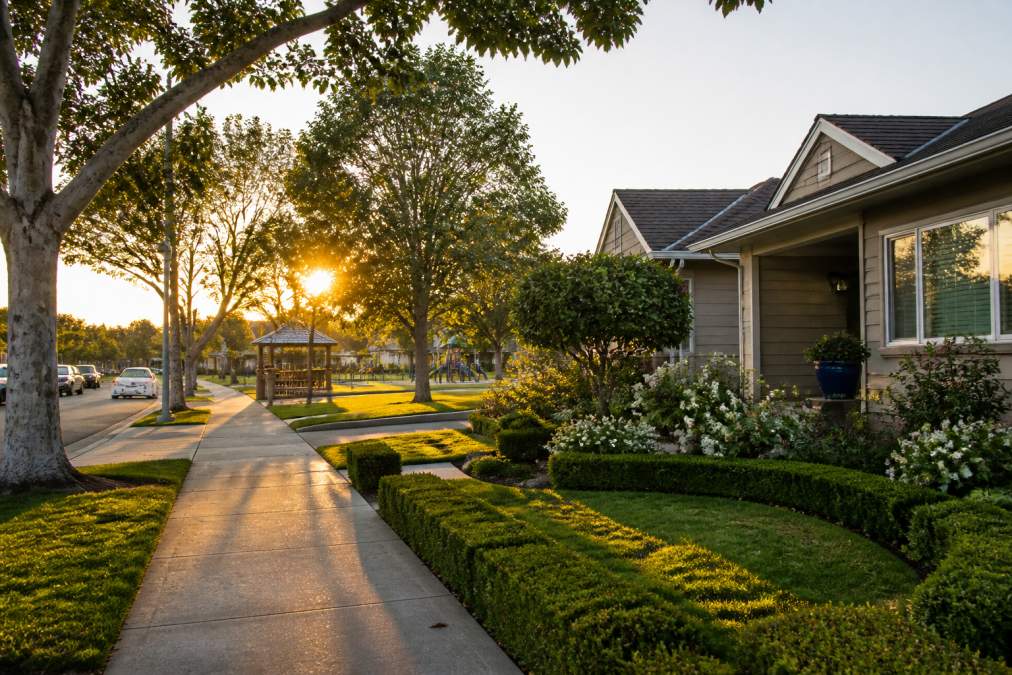 Tree-lined residential street in Burbank Rancho with well-maintained homes, wide sidewalks, and a community park visible in the background during golden hour lighting
