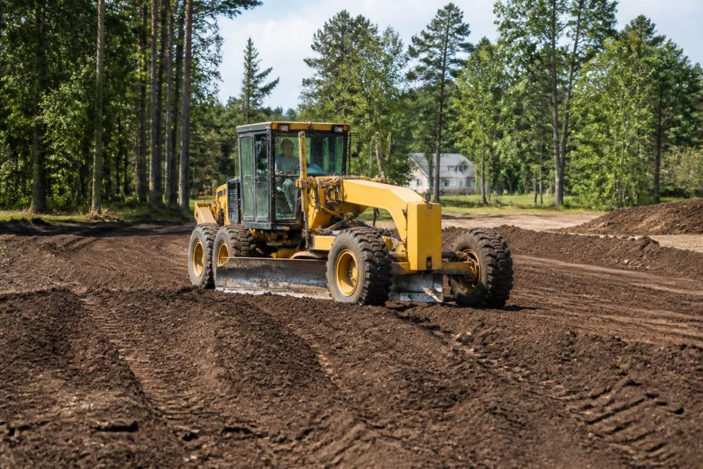 Heavy equipment operator using a grader to shape and level residential property terrain, with fresh soil visible and established trees in background