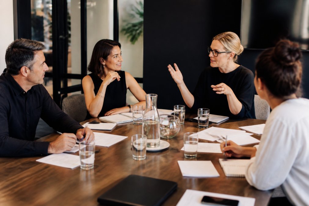 A small group of professional founders and consultants engaged in a live mentoring session around a modern conference table, laptops open and notes spread across the surface, warm overhead lighting creating an engaged, focused atmosphere