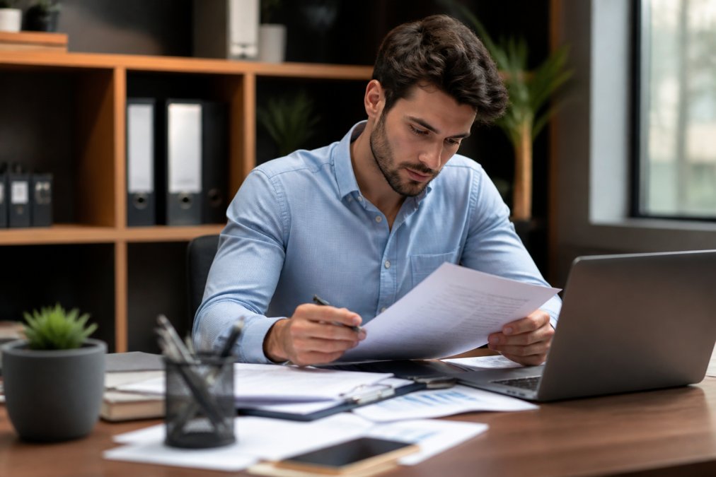 Young entrepreneur at desk reviewing business documents and insurance paperwork with laptop open, professional office setting