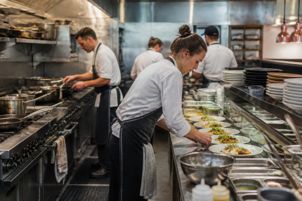 Busy kitchen scene with multiple staff members actively preparing food during peak service hours, with stainless steel equipment, hot stations, and organized food stations visible under bright overhead lighting