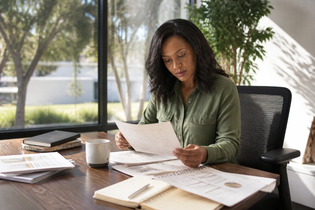 Business owner reviewing strategic planning documents and workbooks at desk with laptop, notebook, and coffee in modern office setting with natural window lighting