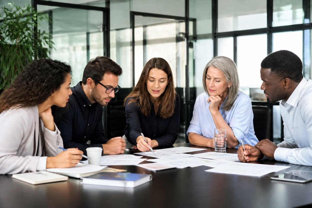 Diverse management team collaborating around modern conference table during business strategy session, reviewing documents with engaged expressions and natural office lighting
