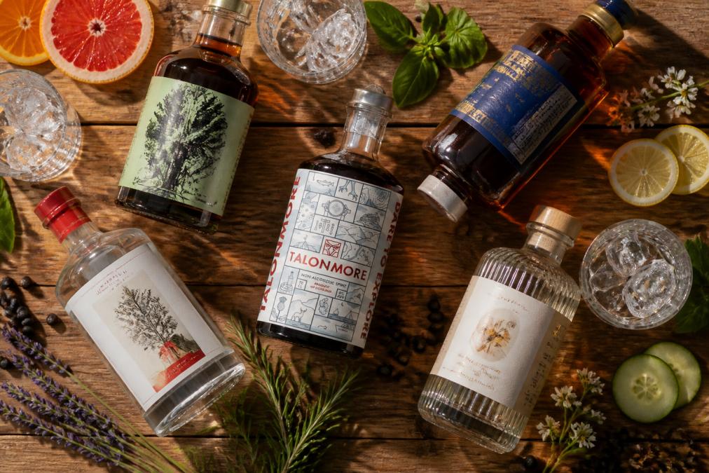 Overhead shot of various 0% alcohol spirit bottles arranged on a wooden table with fresh botanicals, citrus slices, and ice glasses ready for serving