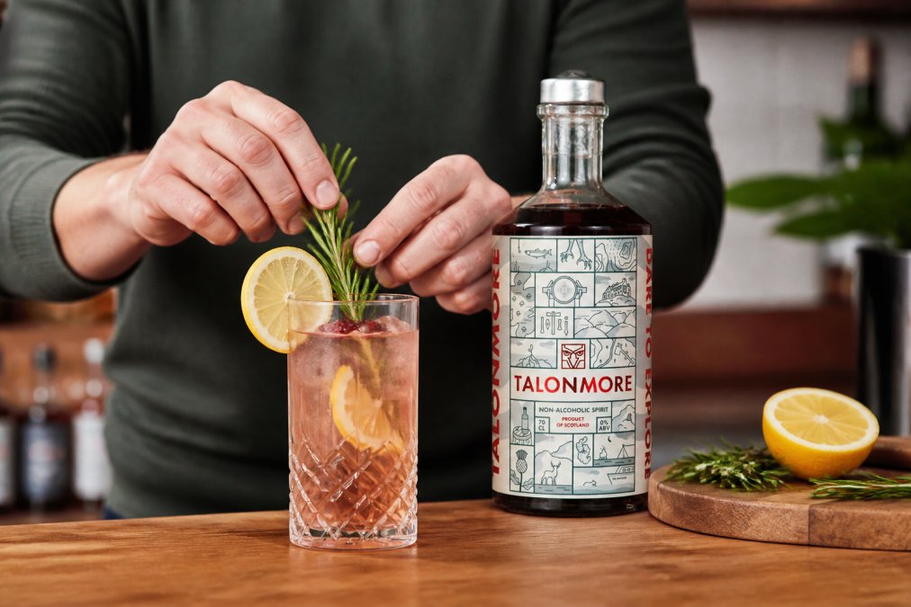 Close-up of hands preparing a colourful alcohol-free cocktail in a glass with fresh herbs, ice, and a citrus garnish in a home bar setting