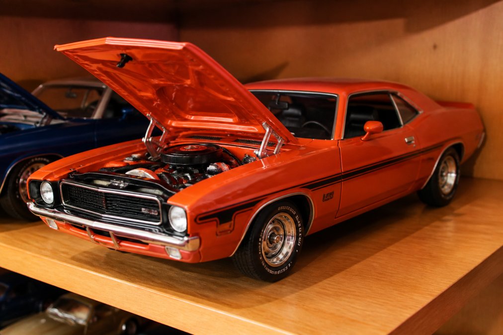 Close-up of a highly detailed 1:18 scale diecast American muscle car on a wooden display shelf, showing open hood with visible engine detail, rubber tires, and glossy paint finish under warm incandescent lighting