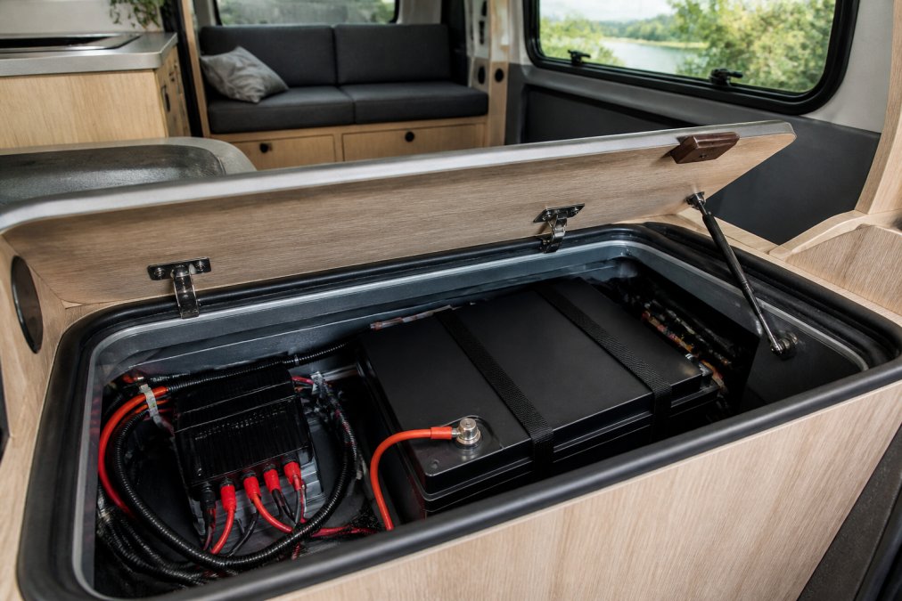Close-up of a 12V LiFePO4 lithium leisure battery installed inside a campervan battery compartment, with wiring and a BMS unit visible and natural daylight coming through a side window
