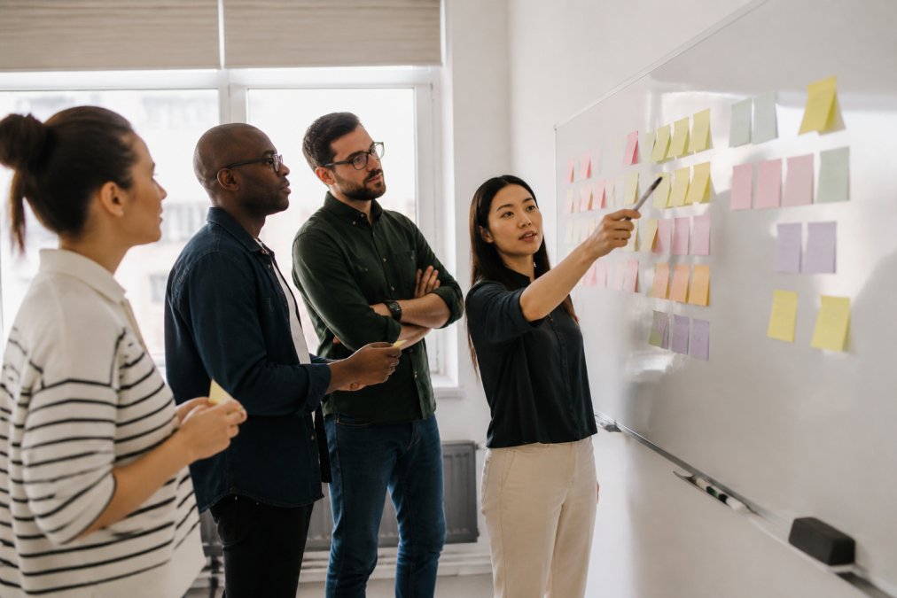 A small agile team of four professionals gathered around a whiteboard covered in sticky notes during a sprint planning session, with natural light from office windows illuminating the group as they point at and discuss the board