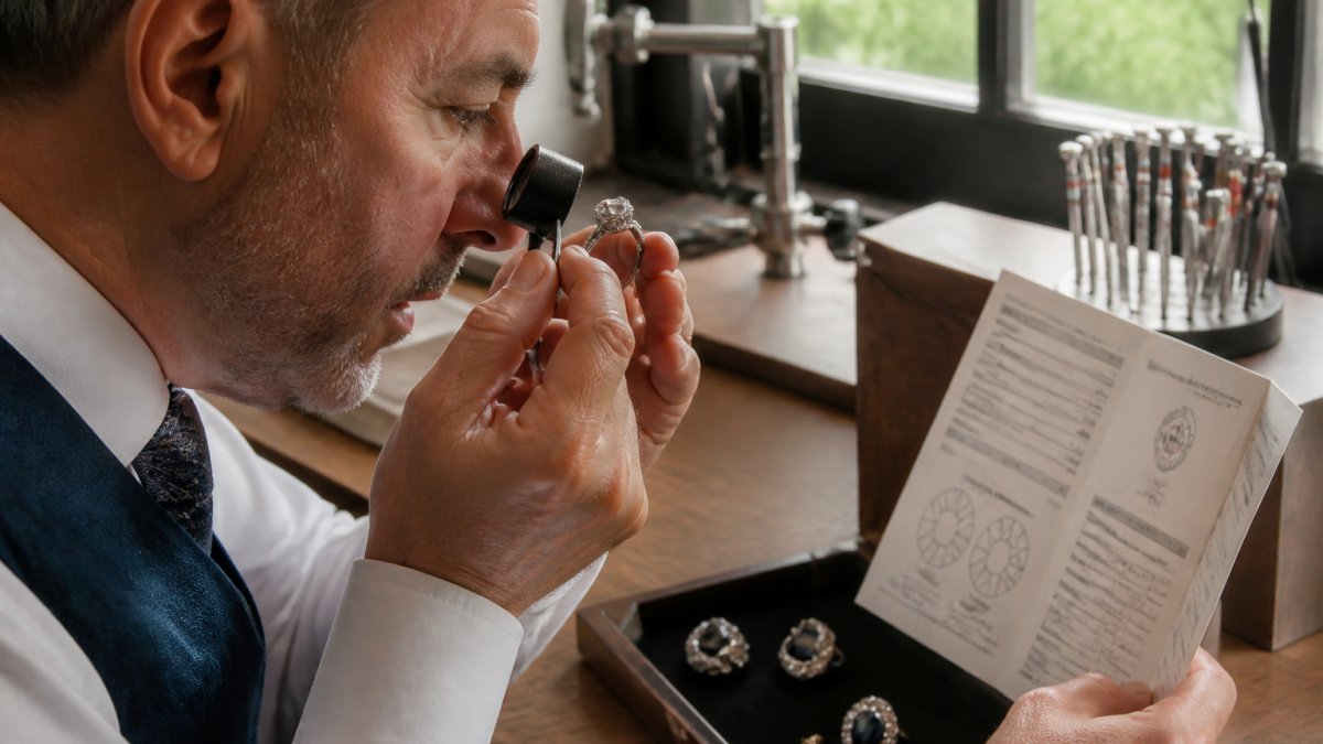 Professional jeweller examining antique diamond ring under magnification with natural light, holding gemological report document, showing facets and hallmarks