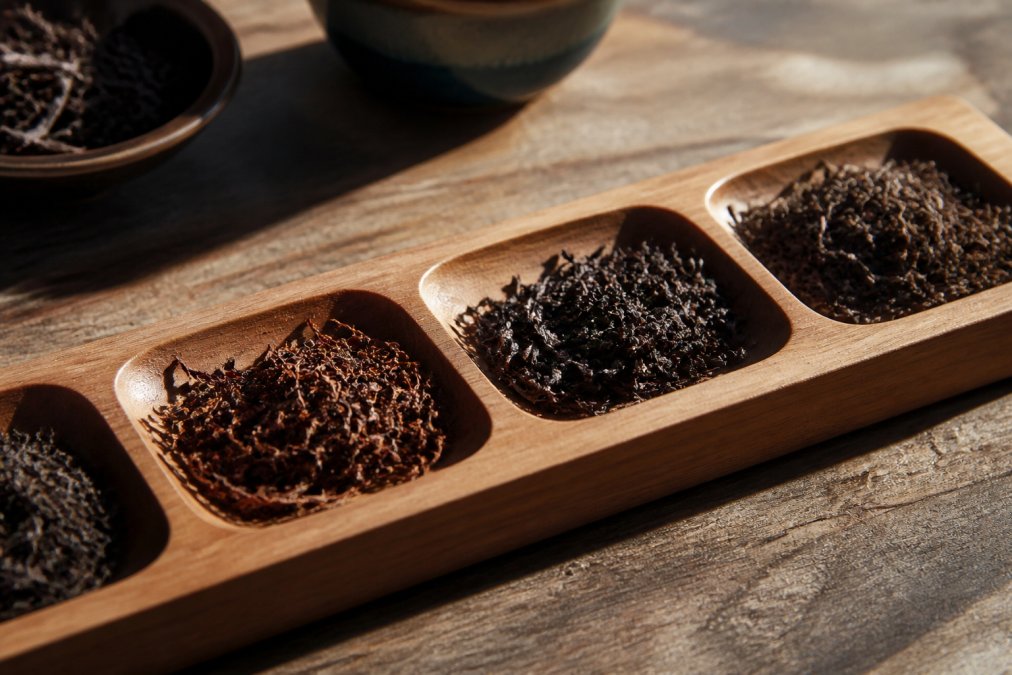 Close-up of loose leaf black tea leaves in various stages of oxidation, showing deep burgundy and black colors in a wooden tasting flight with natural light