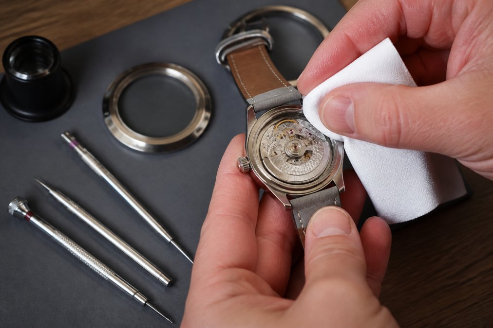 Person carefully cleaning an automatic watch movement with a soft microfiber cloth and specialized watchmaking tools on a clean workbench, showing proper maintenance technique