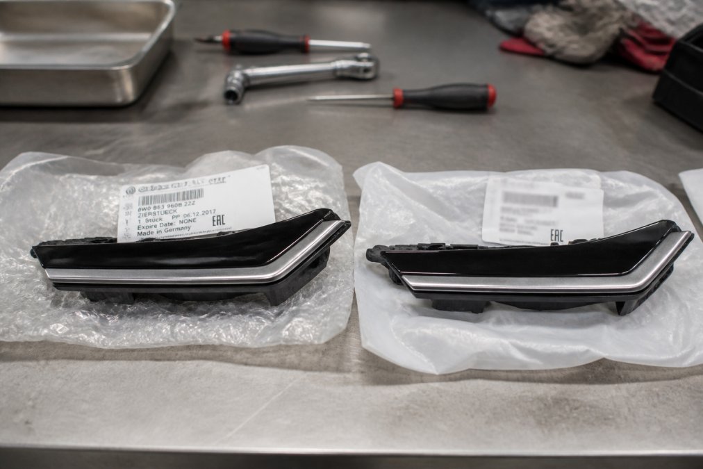 Close-up of two Audi parts side by side on a workbench, one with a clean OEM label and factory packaging, one with a blurry label and cheap plastic bag, under bright garage lighting