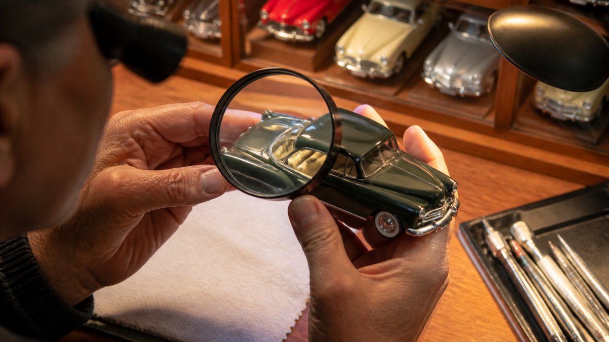 Collector's hands carefully examining a classic diecast car model under warm desk lighting, inspecting the paint finish and interior details with a magnifying glass