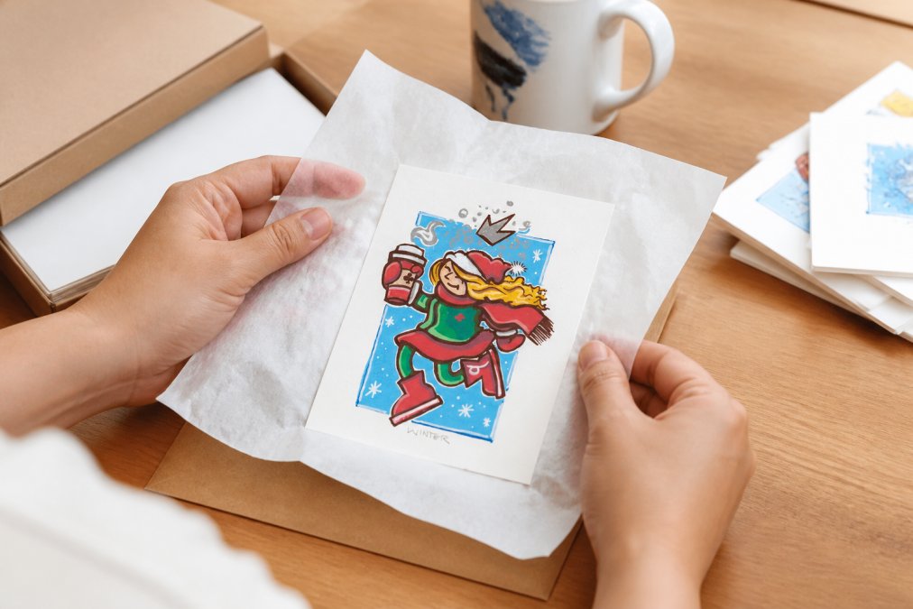 Hands carefully packing a handmade watercolor illustration in protective tissue paper inside a small studio workspace, showing the personal care and attention to detail in each order