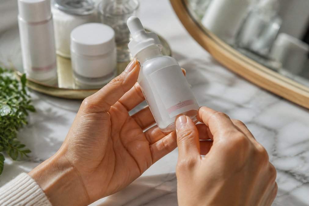 Close-up of hands holding a Korean beauty serum bottle, examining the batch code label and packaging details under natural light, with a marble surface backdrop