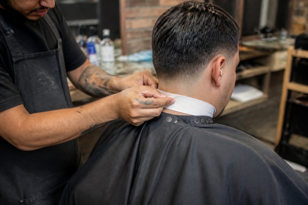 Barber applying a self-adhesive neck strip to a client's neck during an active haircut, demonstrating proper placement around the nape area and the protective barrier in action with professional barbershop lighting