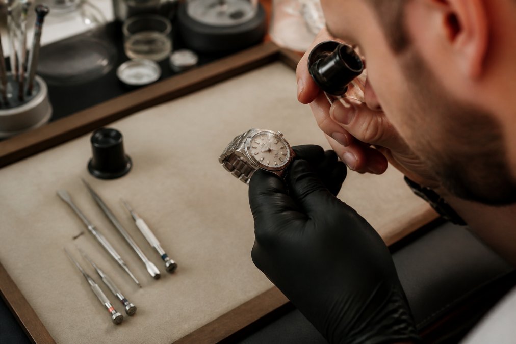 Jeweler using a loupe to inspect the dial and serial number of a certified pre-owned Rolex watch at a workbench.