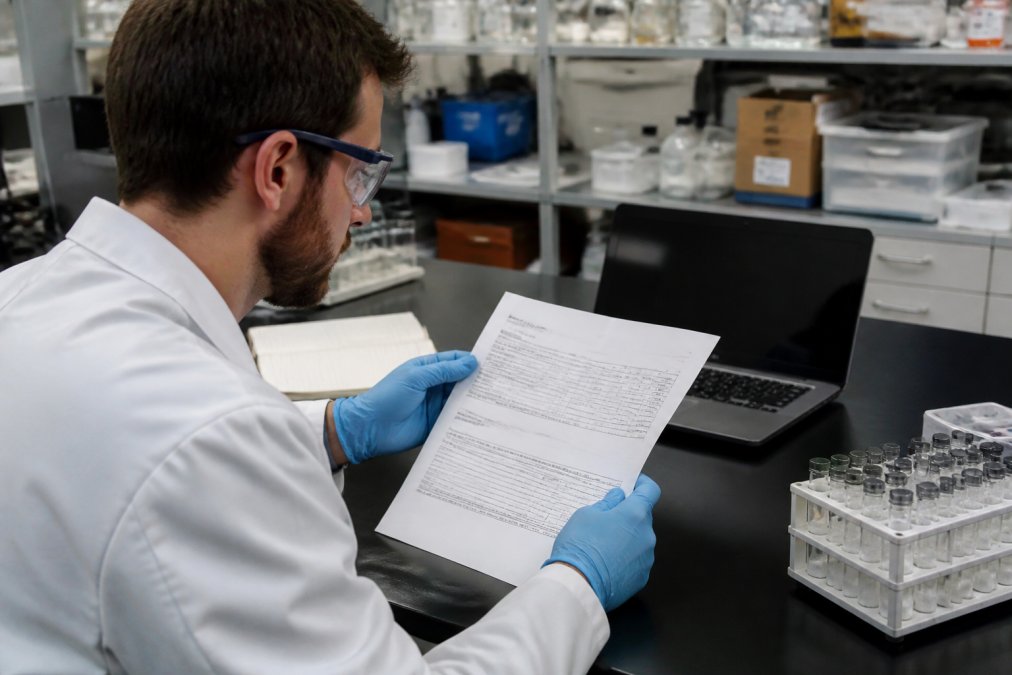 A scientist in a white lab coat and safety glasses reviewing a printed certificate of analysis at a laboratory bench, with a laptop and vial rack visible in the background