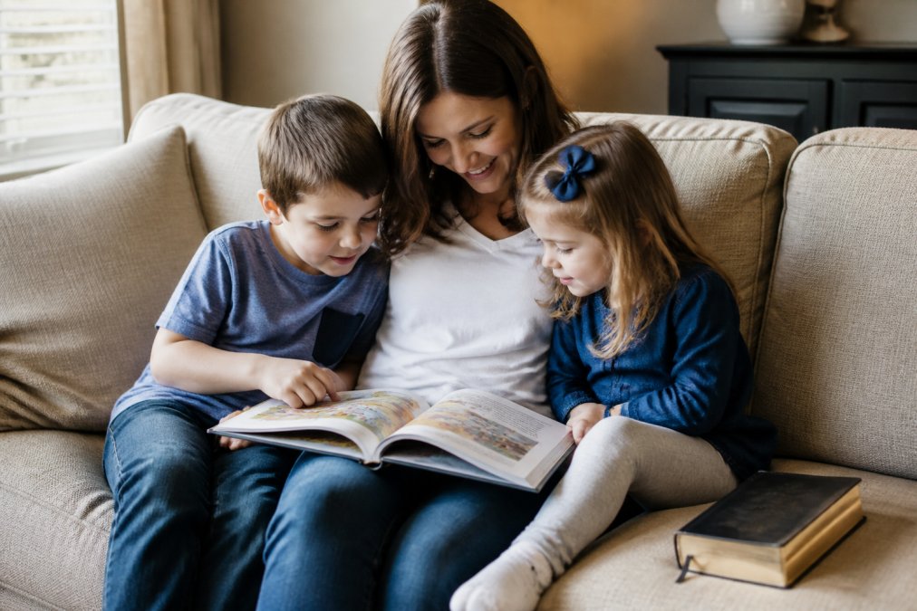 A warm, sunlit living room scene of a mother and two young children sitting on a beige couch, looking down at an open activity book with colorful illustrations, a small Bible resting on the cushion beside them