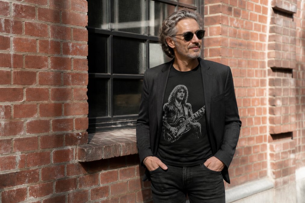 Person in their 50s wearing a classic rock graphic tee styled with dark jeans and a casual blazer, standing confidently in natural lighting with subtle smile