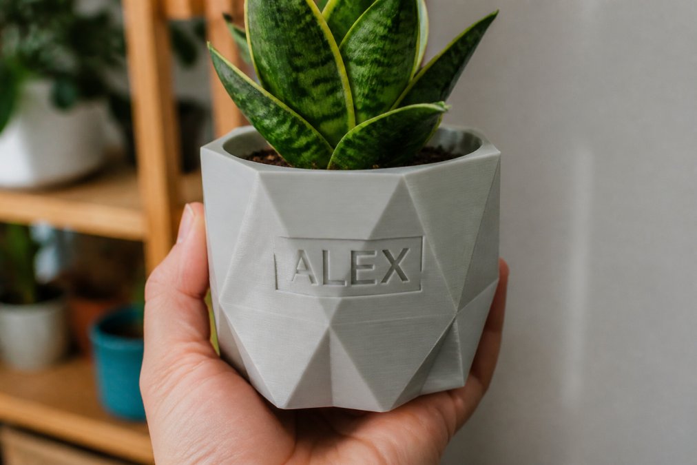 Close-up of a hand holding a custom 3D printed planter with personalized lettering etched into the surface, showing geometric design details and smooth surface finish quality in natural lighting