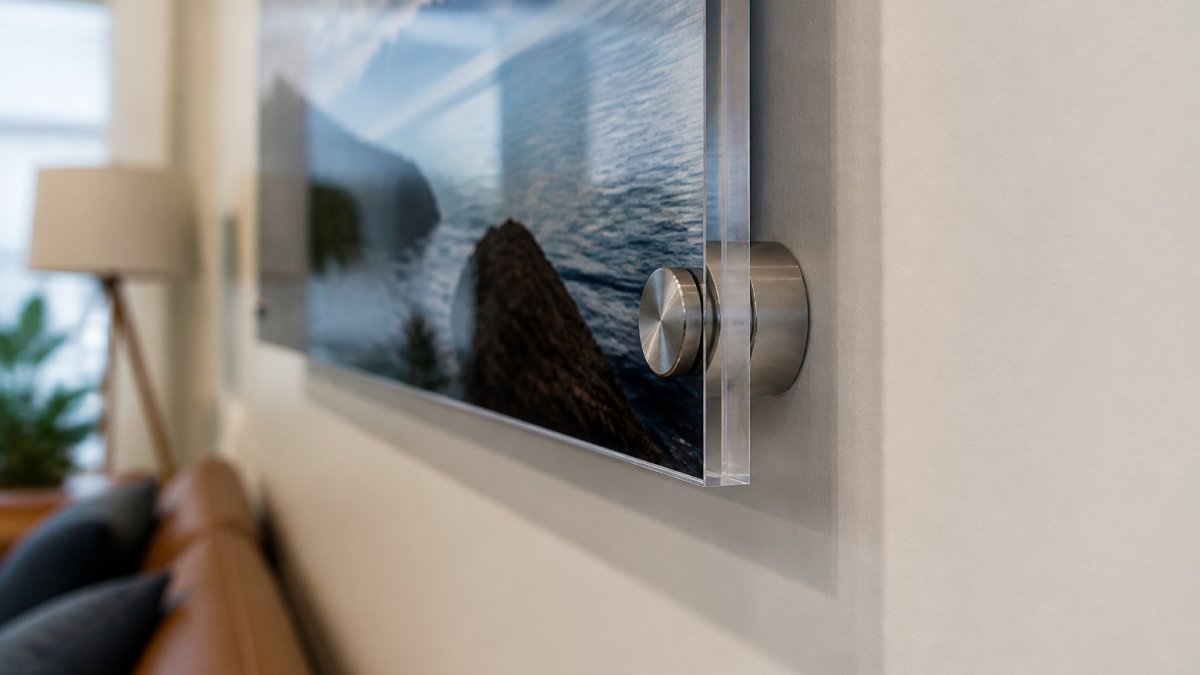 Close-up of polished acrylic edge with stainless steel standoff mounting hardware installed on a modern living room wall, showing the floating effect with subtle shadow beneath the artwork
