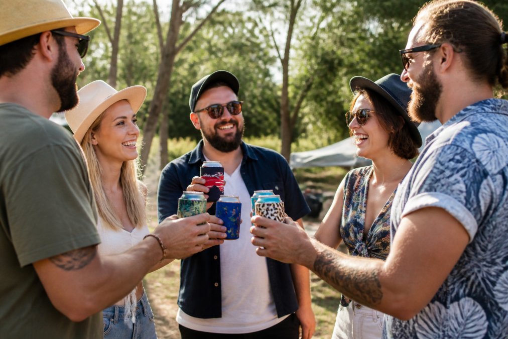 Group of friends at a casual outdoor gathering holding beverages in custom-printed neoprene stubby holders with vibrant designs showing different patterns and colours, the holders visible in real use during daytime
