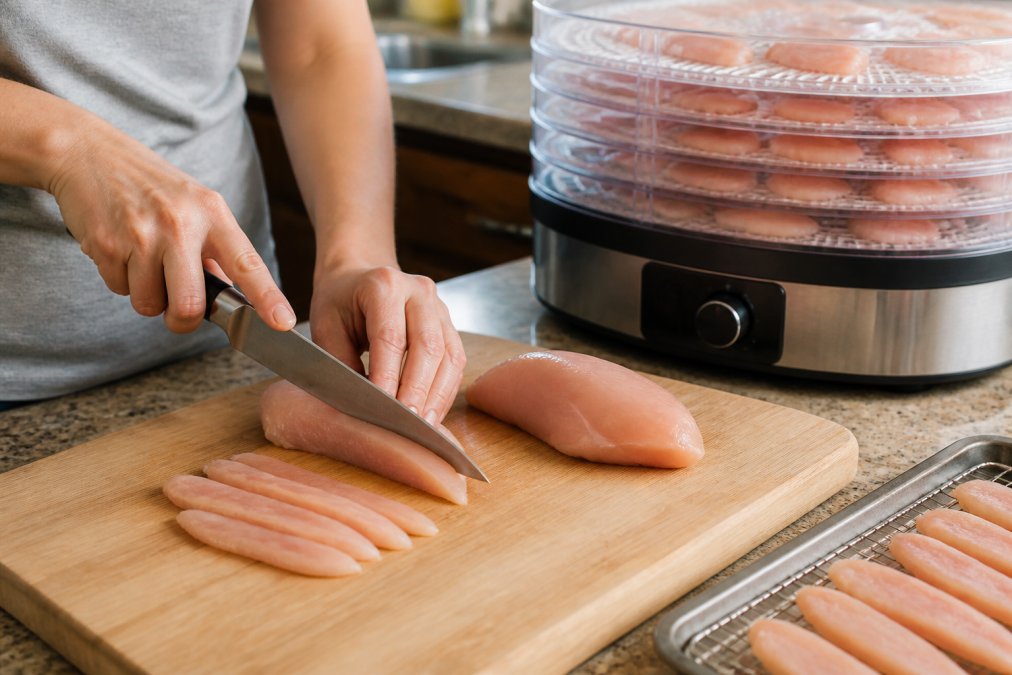 Close-up of a person's hands slicing raw chicken breast into thin strips on a wooden cutting board in a home kitchen, with a food dehydrator visible in the background