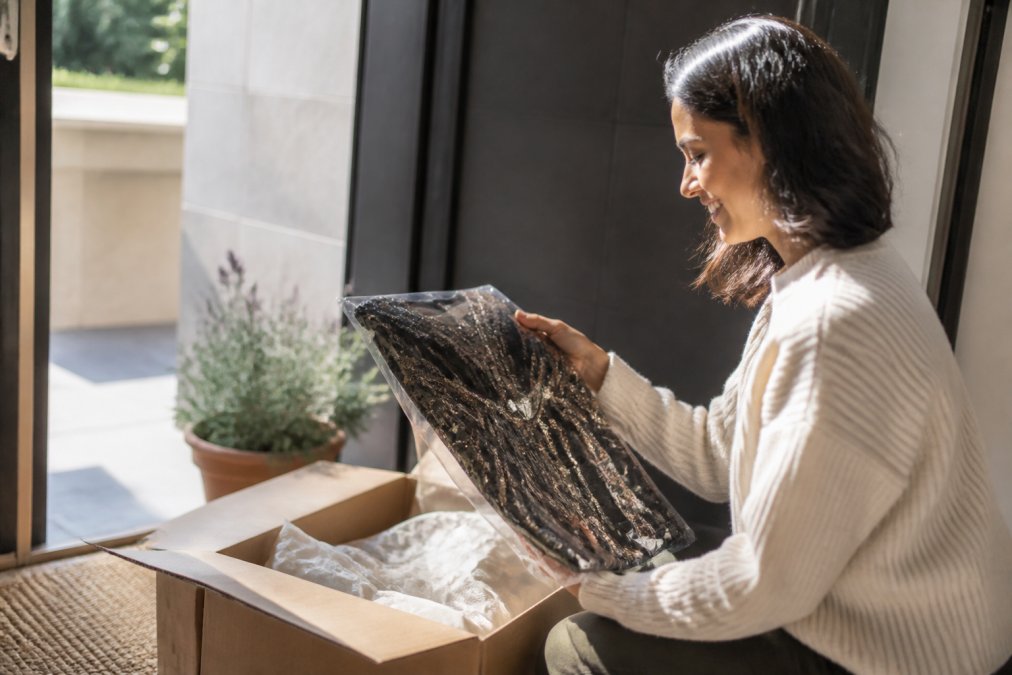 Woman unpacking a carefully packaged designer dress delivery at her doorstep, examining the quality of packaging and garment with morning light