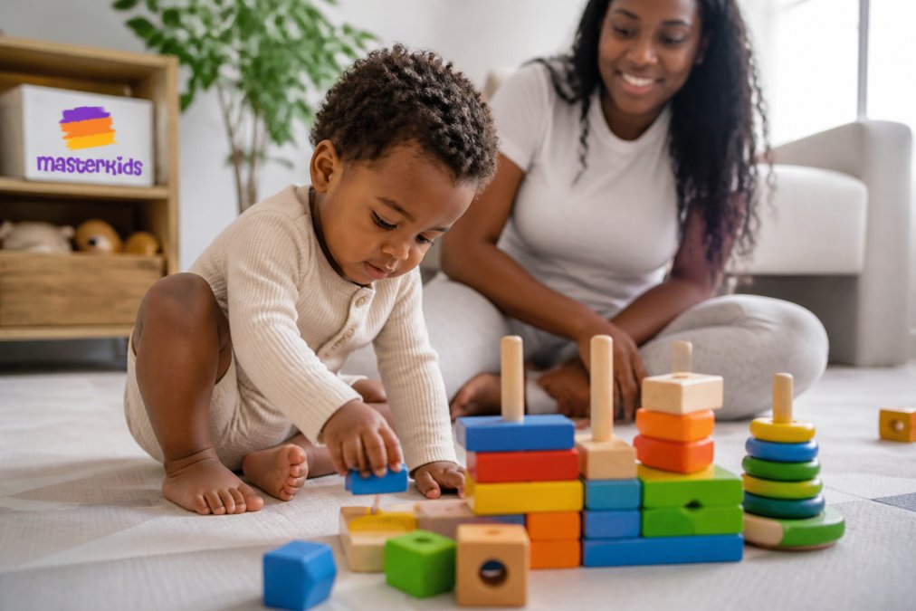 Young child playing with colourful wooden blocks and stacking toys on a clean playmat, parent supervising in background