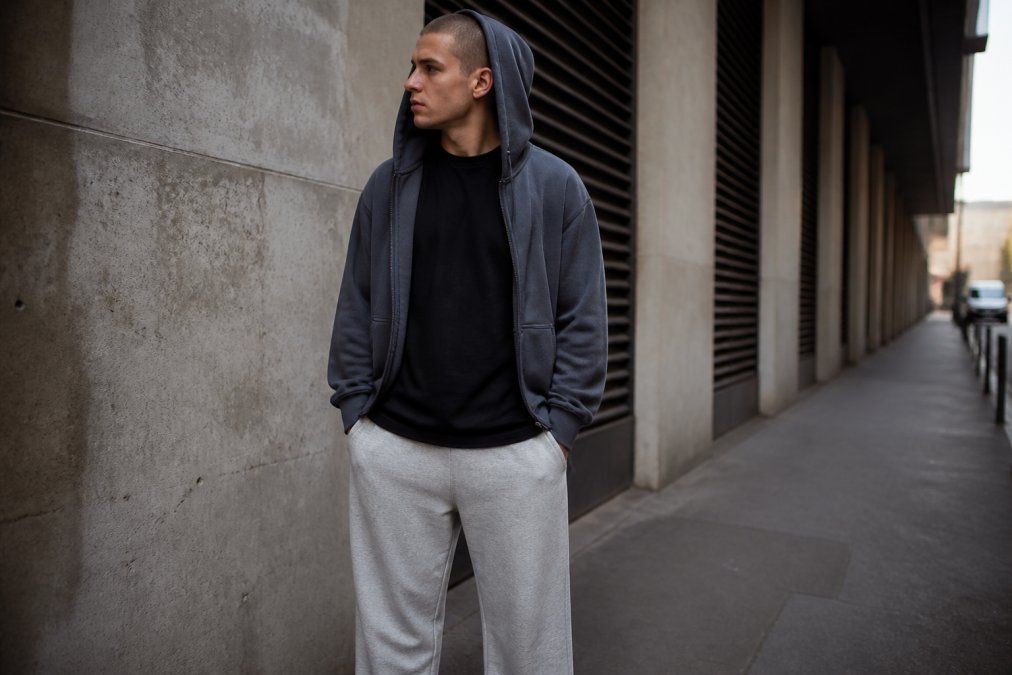 Man wearing oversized heavyweight black tee layered under a slate-colored hoodie, paired with relaxed-fit sweatpants in neutral tone, styled in an urban setting with natural daylight