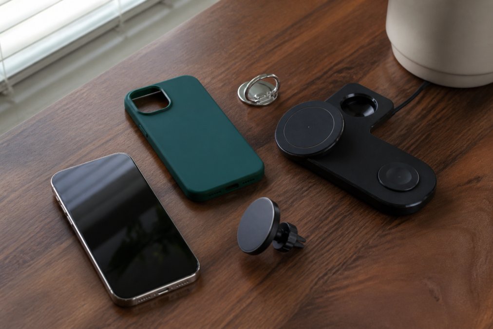 Flat-lay photo on a walnut desk showing a soft silicone phone case in emerald, a silver metal ring stand, a three-in-one wireless charger, and a magnetic car phone holder arranged beside a smartphone, soft window light from the left