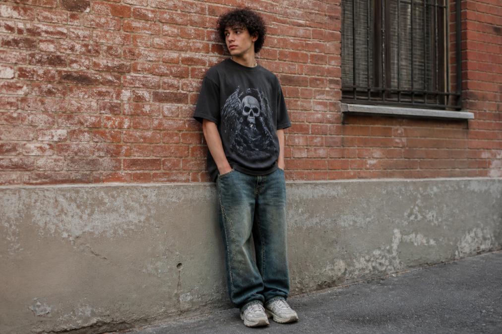 Young adult wearing an oversized vintage graphic tee with loose jeans and chunky sneakers against a brick wall