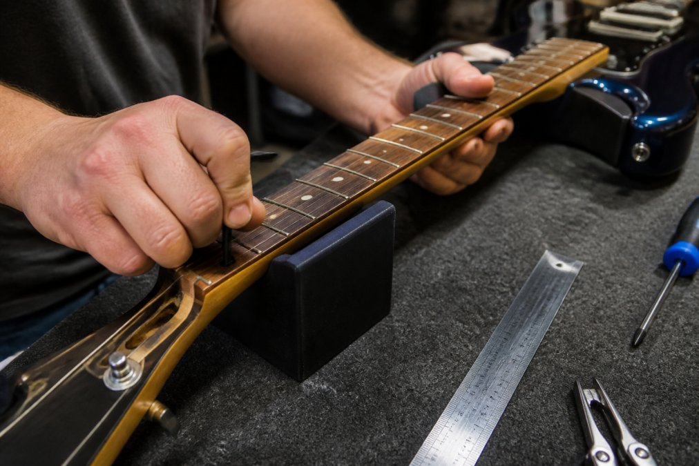 Hands adjusting the truss rod on an electric guitar neck with an Allen wrench on a workbench, with a measuring ruler and other setup tools visible nearby