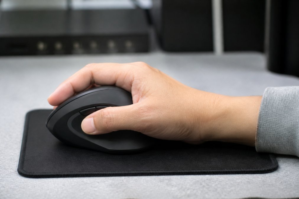 Close-up of hand holding ergonomic mouse with wrist in neutral position, fingers naturally curved around contoured side, sitting on mousepad at desk