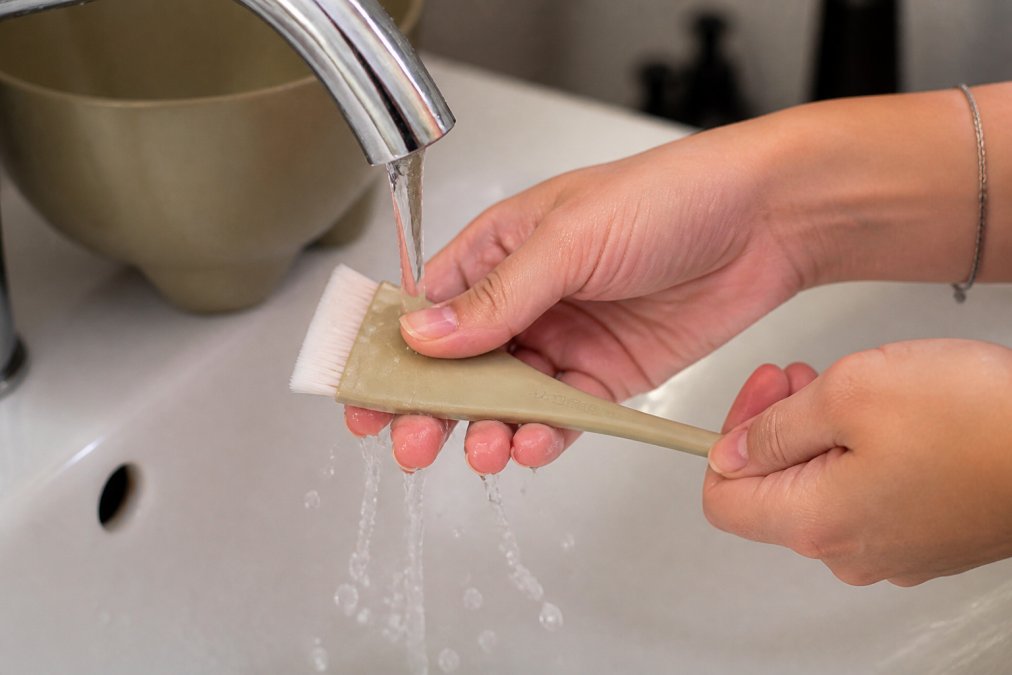 Close-up of hands rinsing professional hair color brush under running water with proper cleaning technique in bright bathroom lighting