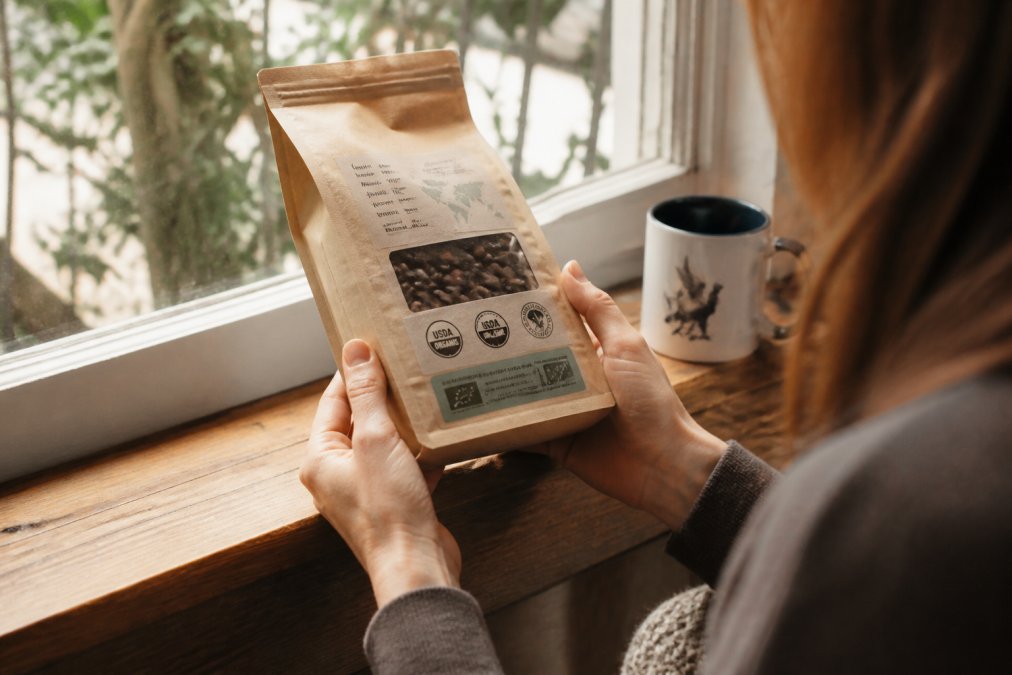 Person's hands holding a bag of fresh roasted coffee beans, examining visible certification labels and origin information printed on kraft packaging in natural window light