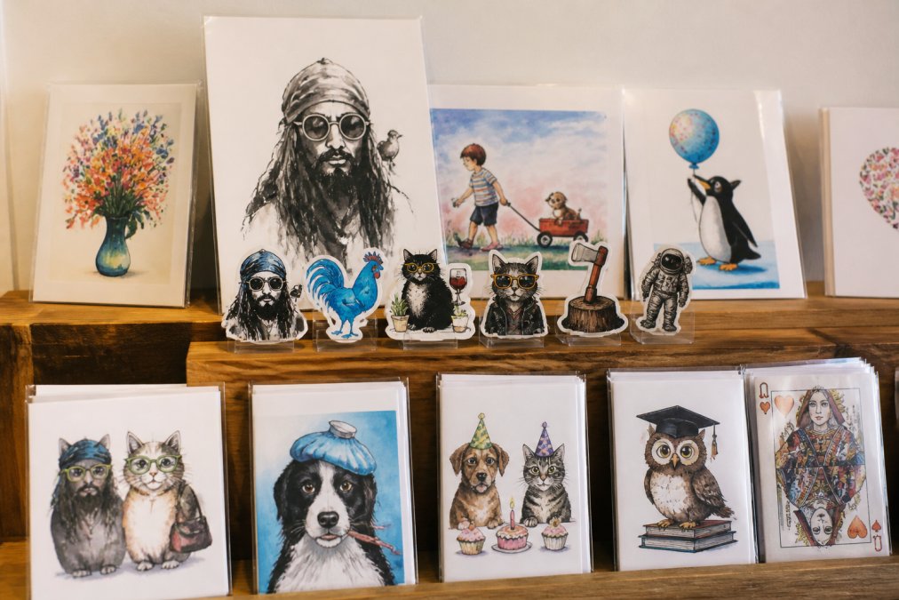 A collection of handmade original art pieces and vinyl stickers displayed together on a wooden shelf, showing variety of artistic styles, watercolor textures, and ink illustrations arranged with genuine artistic presentation